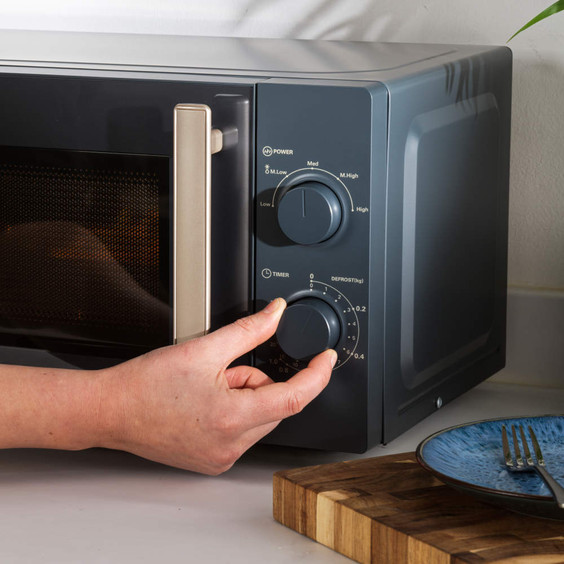 Grey microwave oven on kitchen counter, hand adjusting timer dial, plate and fork nearby