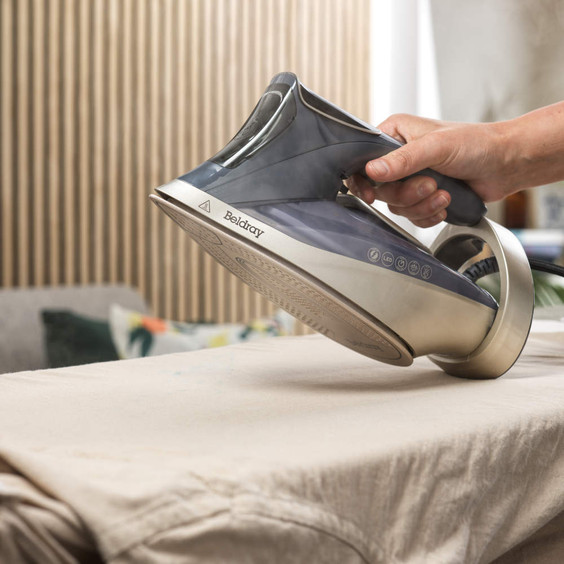 Hand holding gold steam iron on beige shirt, in use for ironing clothes.