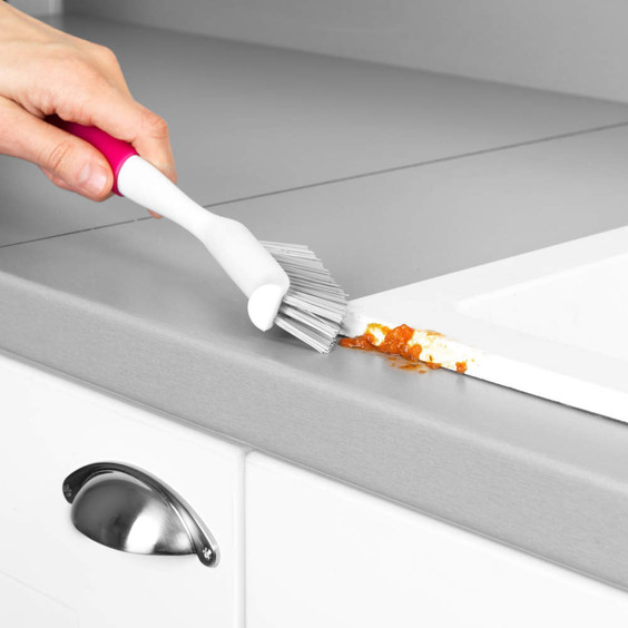 Narrow cleaning brush with pink handle scrubbing food residue from kitchen worktop edge