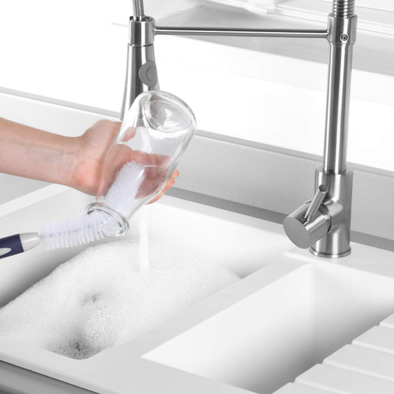 Bottle brush cleaning a glass bottle under running water in a white kitchen sink.