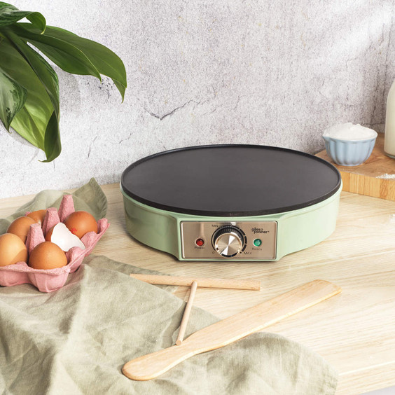 Green electric crepe maker on worktop with eggs, flour and milk, shown for kitchen product listing.