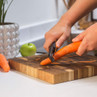 Hands peeling a carrot with a black peeler on a wooden chopping board in a kitchen