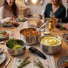 Stainless steel cookware set in use at a family dinner table, featuring stew and vegetables.