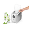Countertop ice maker in stainless steel on garden table with summer drinks and fruit.
