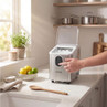 Silver countertop ice maker with open lid, showing ice cubes and a hand pressing controls