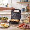 Black sandwich toaster making two toasted sandwiches with salad and toppings nearby