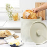 Cream bread bin with ribbed sides and lid, hand placing loaf inside, shown in kitchen