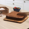 Square chocolate cake being sliced on a wooden board in a kitchen, with baking tin nearby