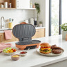 Orange electric grill open on kitchen counter, burger and patties beside, salad and sauces nearby