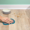 Hand wiping pet crumbs from wooden floor near dog bowl and pink pet bed