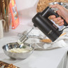 Black hand mixer with gold controls in use, beating cream in a metal bowl on a kitchen counter