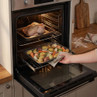 Roasting tray with assorted potatoes and carrots being placed in oven beside whole chicken