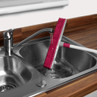 Pink floor brush with white handle being rinsed under tap in stainless steel kitchen sink
