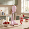 Pink milkshake maker with metal jug on kitchen counter, next to strawberry milkshake