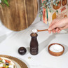 Dark wood salt grinder on kitchen worktop, hand filling with salt using wooden spoon