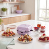 Purple waffle maker on kitchen counter with plates of waffles, fruit, and syrup
