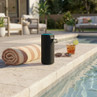 Black portable Bluetooth speaker by poolside, shown with towel and drink