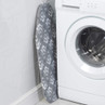 Grey ironing board with white floral pattern standing upright beside washing machine