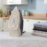 Electric steam iron upright on ironing board, folded shirts beside, kitchen background