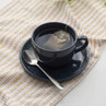 Black cup and saucer with hot tea bag and teaspoon on patterned cloth