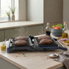 Open electric grill cooking four beef burgers in kitchen, ingredients prepared beside.