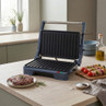 Blue electric grill press open on kitchen counter, steak and kebabs beside