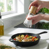 Clear salt grinder with wooden top in use, seasoning chicken and vegetables in cast iron pan