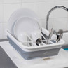 Grey and white collapsible dish drainer with clean plates, mugs, and cutlery beside sink