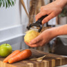 Black-handled peeler in use on potato, carrots and green apple on chopping board.