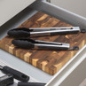 Pair of stainless steel kitchen tongs with black tips on wooden chopping board in drawer