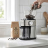 Electric coffee grinder in brushed steel. Whole beans being poured in. Kitchen scene.
