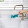Blue kitchen scrub brush being rinsed under a tap over a white sink