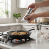 Silver pepper grinder seasoning chicken in skillet on gas hob, salt grinder visible