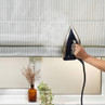 Hand holding steam iron next to blue striped roman blind over window, lifestyle setting