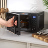 Black digital microwave oven with blue LED display, hand turning control knob, on kitchen counter