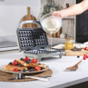 Grey waffle maker in kitchen, batter being poured in, finished waffles with berries on plate