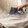 Hand using Beldray steam iron on beige fabric, steam visibly removing creases