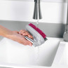 Hand rinsing a grey scrubbing brush with pink bristles under a kitchen tap