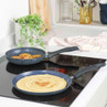Set of two non-stick frying pans on induction hob, one with pancakes, one with apple slices.