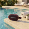 Red wireless speaker on poolside table beside towel and plant in sunny outdoor setting