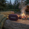Portable red Bluetooth speaker on log by campfire, outdoor camping setting