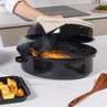 Black enamel roasting pan with lid, shown with cooked chicken and herbs inside on kitchen worktop.