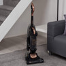 Upright vacuum cleaner in rose gold and black, shown on carpet beside sofa and stairs