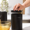 Black metal sugar canister with lid being lifted, next to coffee jar and mugs on kitchen counter