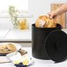 Black ribbed bread bin with lid, hand placing round loaf inside on kitchen worktop