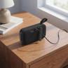 Black portable Bluetooth speaker charging on bedside table, rear view showing cable.