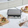 Stainless steel contact grill on kitchen counter, lid closed, with sandwiches and kebabs nearby