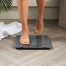 Person stepping onto black glass digital bathroom scale on wood floor