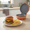 Orange electric grill open beside plate with cheeseburger and chips on kitchen worktop