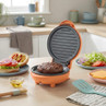 Orange burger maker with cooked patty, next to prepared bun and salad toppings on kitchen counter